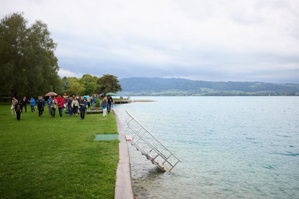 Menschen laufen am Ufer eines Sees entlang, während einige Regenschirme halten. Im Hintergrund sind Bäume und Hügel sichtbar.