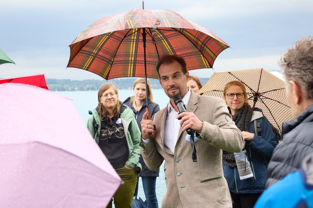 Eine Gruppe von Personen steht am Wasser, während ein Mann mit Mikrofon in der Hand spricht und einen karierten Regenschirm hält. Weitere Teilnehmer, die ebenfalls Regenschirme halten, hören aufmerksam zu.