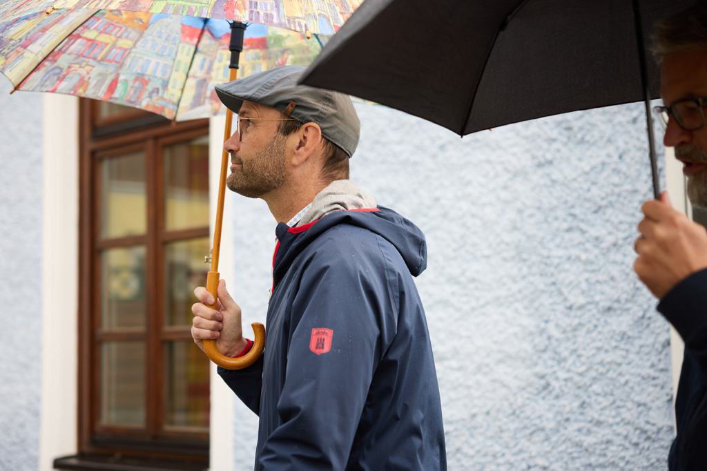 Ein Mann mit einer bunten Regenschirm und einer blauen Jacke geht entlang einer blauen Wand, während ein anderer Mann mit einem schwarzen Regenschirm hinter ihm steht.