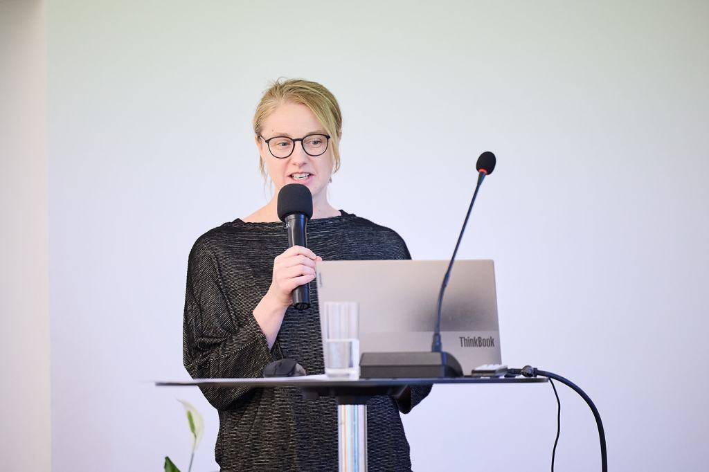 Frau mit Brille spricht am Mikrofon vor einem Laptop auf einem Tisch.