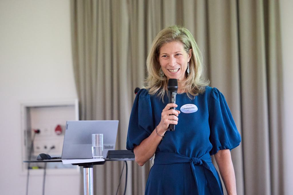 Eine lächelnde Frau mit blonden Haaren hält ein Mikrofon und spricht in ein Publikum. Sie trägt ein blaues Kleid und steht vor einem Tisch mit einem Laptop und einem Glas Wasser.