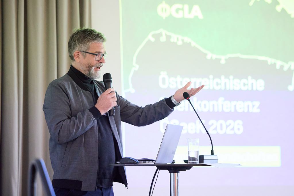 Ein Mensch mit Brille spricht und präsentiert auf einer Konferenz, im Hintergrund ist ein Bildschirm mit Informationen zur österreichischen Geoinformationskonferenz 2026 zu sehen.