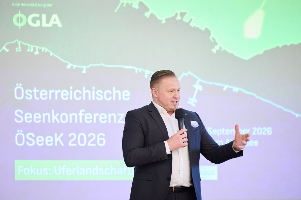 Ein Mann spricht auf einer Bühne während der Österreichischen Seen-Konferenz ÖSeeK 2026, im Hintergrund ist ein Präsentationsscreen mit Informationen zur Veranstaltung zu sehen.