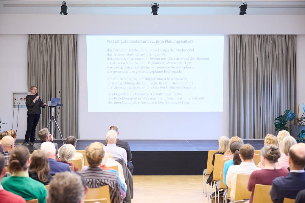 Eine Präsentation über gute Baukultur und Planungskultur, gehalten von einer Frau auf einer Bühne, während das Publikum zuhört. Auf der Leinwand sind Texte zu den Themen nachhaltige Architektur, Bürgerbeteiligung und Stadtentwicklung zu sehen.