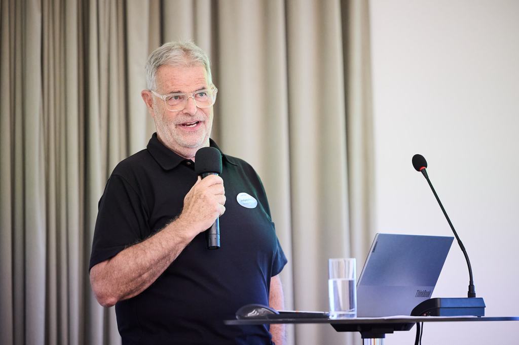 Ein älterer Mann mit Brille hält ein Mikrofon und spricht in ein Publikum. Er trägt ein schwarzes Poloshirt und steht neben einem Tisch mit einem Laptop und einem Glas Wasser. Im Hintergrund sind Vorhänge zu sehen.