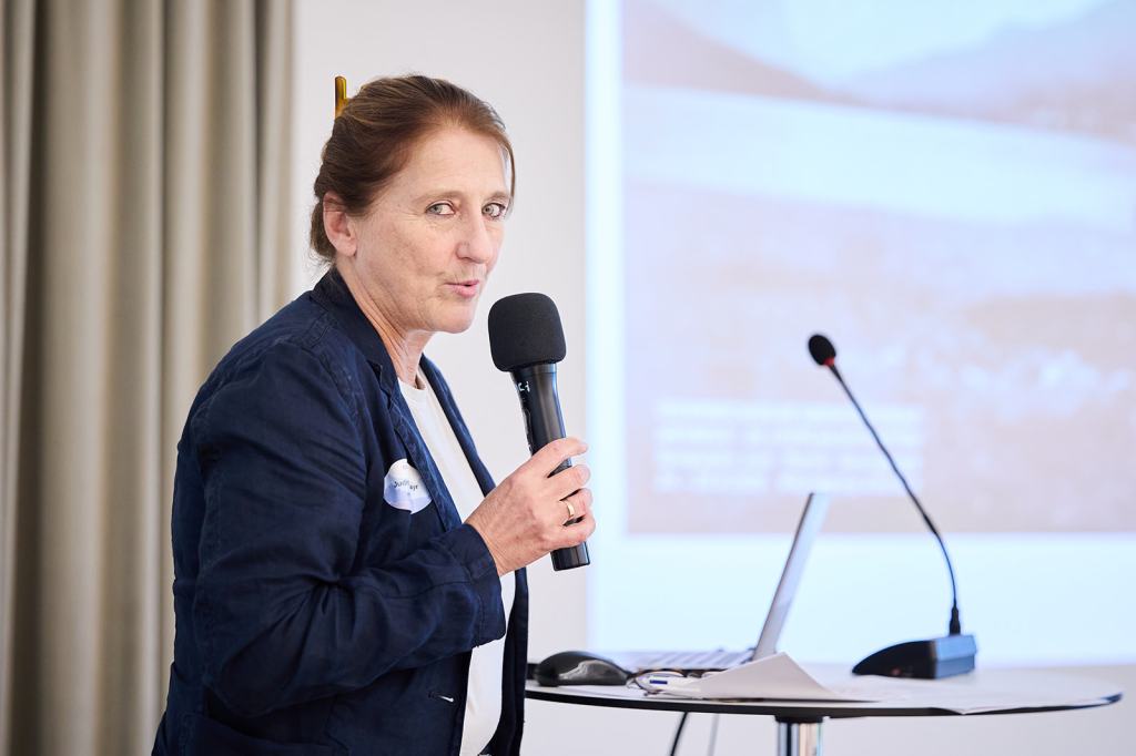 Eine Frau steht an einem Pult und spricht mit einem Mikrofon in der Hand, während im Hintergrund eine Präsentation sichtbar ist.