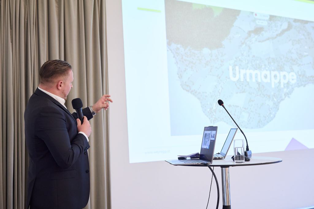 Ein Mann in einem Anzug präsentiert vor einer Folie mit der Aufschrift 'Urmappe', während er einen Zeigestab hält.