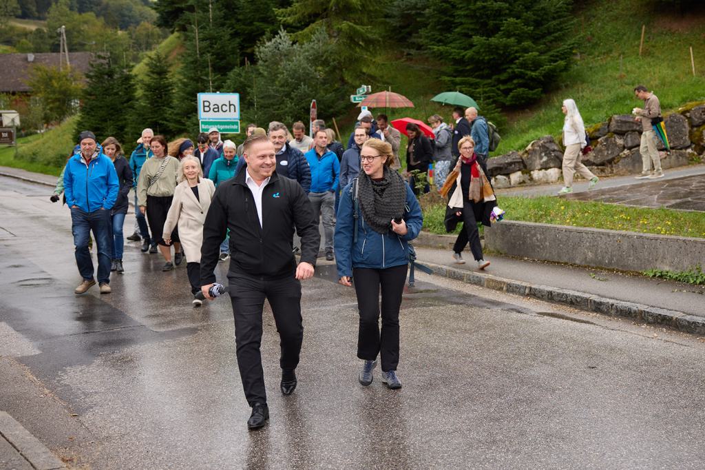 Eine Gruppe von Menschen geht eine Straße entlang, während sie in der Nähe eines Schildes mit der Aufschrift "Bach" spazieren. Einige tragen Regenschirme und die Umgebung ist grün und ländlich.