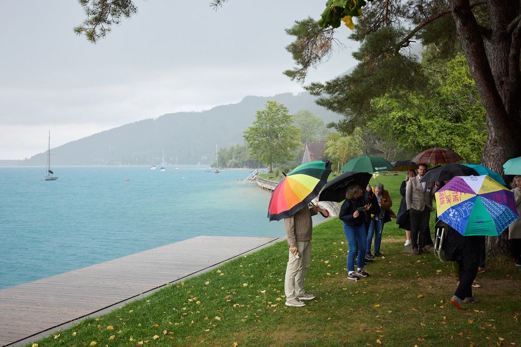 Gruppe von Menschen mit Regenschirmen steht am Ufer eines Sees an einem bewölkten Tag, mit Segelbooten im Hintergrund.