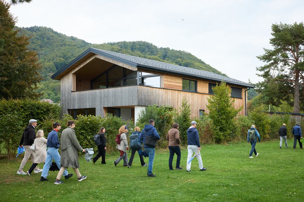 Eine Gruppe von Personen, die auf einer Wiese spazieren gehen, mit einem modernen Holzhaus im Hintergrund und sanften Hügeln im Hintergrund.