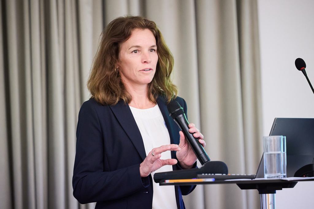 Eine Frau mit lockigem Haar trägt einen Anzug und spricht mit einem Mikrofon in der Hand vor einem Publikum, während sie an einem Podium steht.