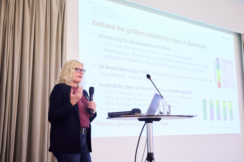 Eine Präsentation mit einer redenden Person, die Informationen über den Zustand natürlicher Seen in Österreich teilt. Im Hintergrund sind Folien mit Daten zur Umsetzung der EU-Wasserrahmenrichtlinie und Ergebnisanalysen sichtbar.