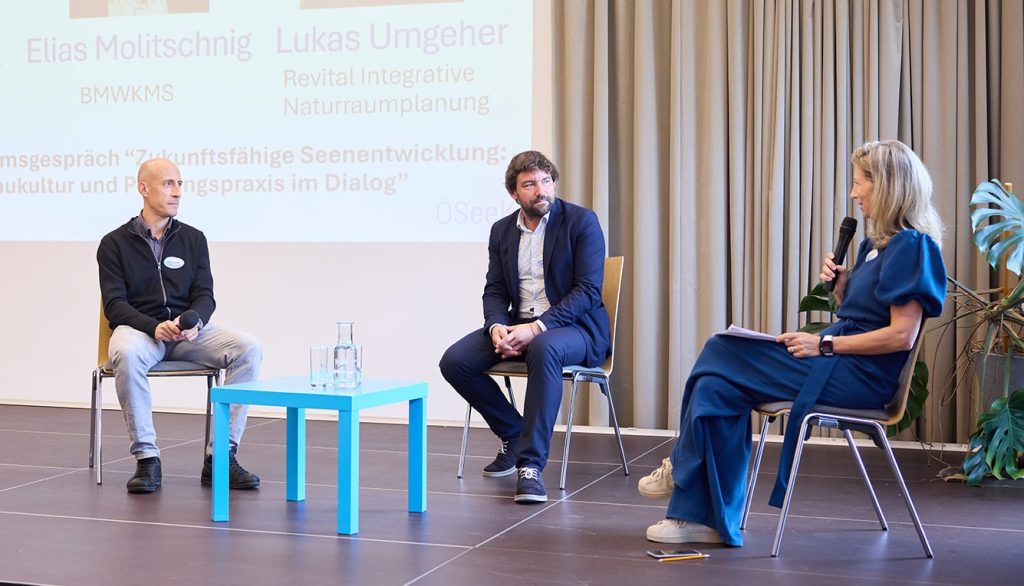 Drei Personen diskutieren auf einer Bühne. Zwei Männer sitzen auf Stühlen, während eine Frau mit einem Mikrofon spricht. Im Hintergrund sind Vortragsunterlagen und ein Pflanzenarrangement zu sehen. Das Thema der Diskussion ist 'Zukunftsfähige Seentwicklung'.