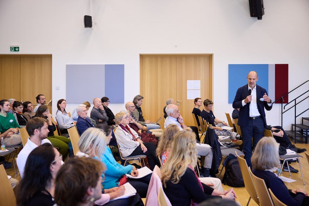 Eine Szene von einer Präsentation, bei der ein Referent vor einem Publikum spricht. Das Publikum sitzt in einem großen Raum mit Holzfußboden und ist aufmerksam. Einige Personen nehmen Notizen oder hören aktiv zu.