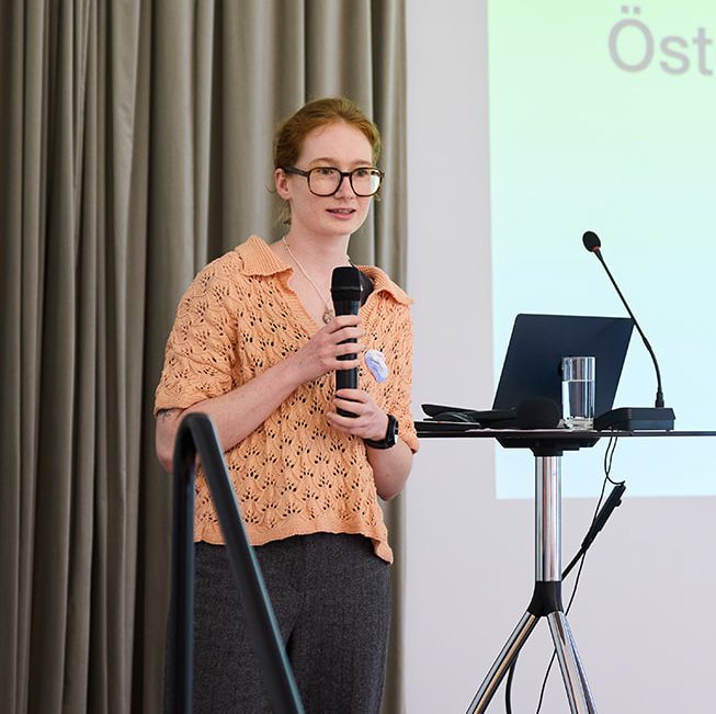 Eine Person steht an einem Rednerpult und hält ein Mikrofon, während sie präsentiert. Im Hintergrund ist ein Bildschirm sichtbar.