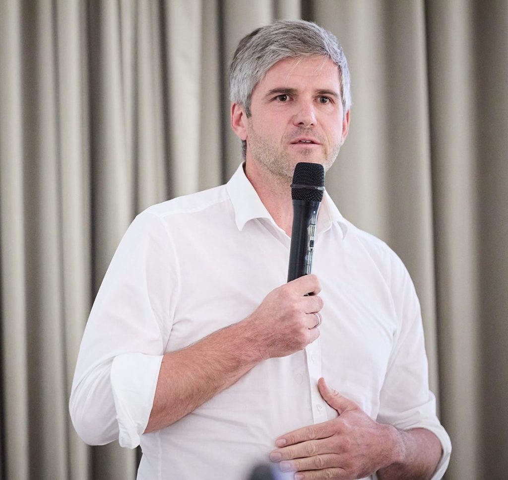 Ein Mann mit grauen Haaren spricht in ein Mikrofon, während er in einem weißen Hemd gekleidet ist. Der Hintergrund besteht aus hellen Vorhängen.