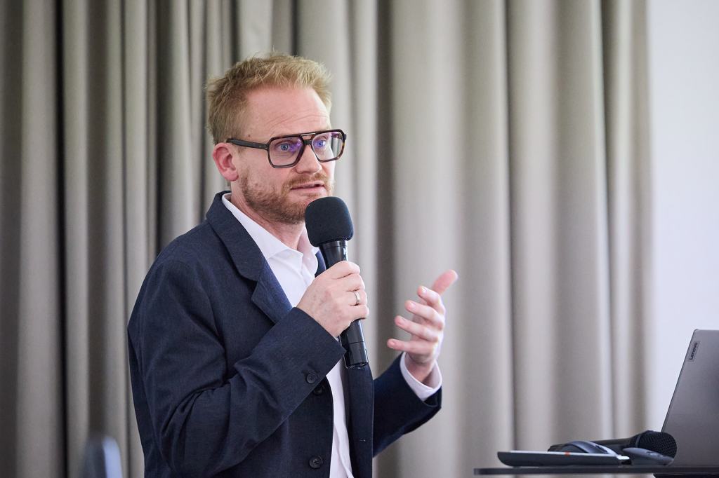Ein Mann spricht mit einem Mikrofon in der Hand, während er eine Präsentation hält. Er trägt eine Brille und hat kurzes, helles Haar. Im Hintergrund sind Vorhänge sichtbar.