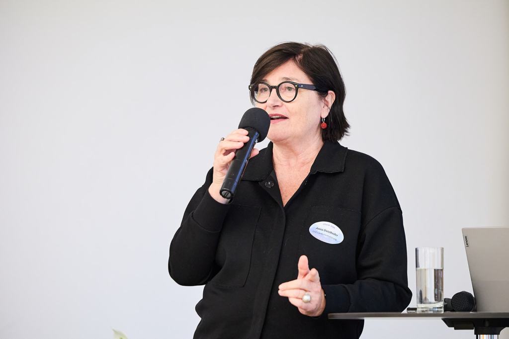 Frau spricht mit einem Mikrofon und ist am Rednerpult, im Hintergrund ein Glas Wasser.