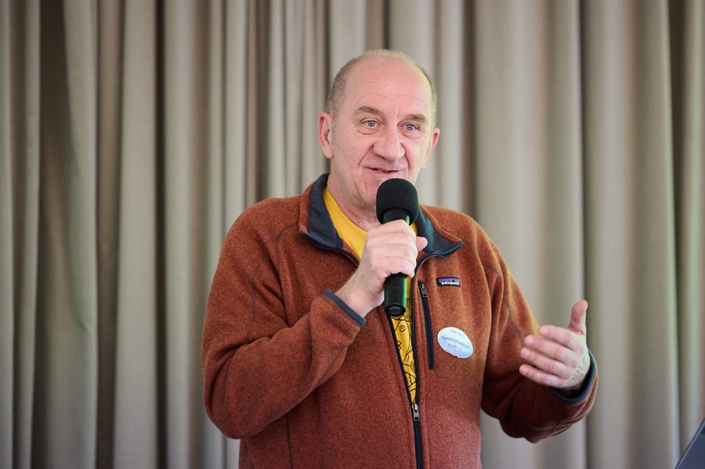 Ein Mann spricht mit einem Mikrofon in der Hand, während er seinen Vortrag hält. Er trägt einen orangefarbenen Pullover und hat eine gelbe T-Shirt darunter. Im Hintergrund sind graue Vorhänge zu sehen.