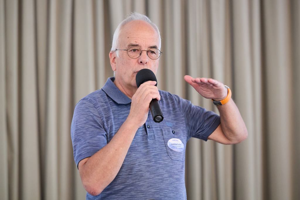 Ein älterer Mann spricht mit einem Mikrofon in der Hand, während er gestikuliert. Er trägt ein blau gestreiftes Polohemd und eine Brille. Hinter ihm ist ein neutraler, hellfarbener Hintergrund.