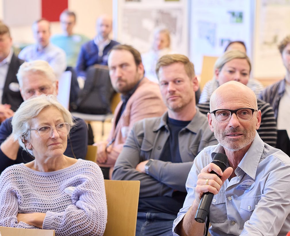 Eine Gruppe von Menschen in einem Raum, die an einer Diskussion teilnehmen. Ein Mann mit Brille spricht in ein Mikrophon, während andere Zuhörer aufmerksam zuhören.