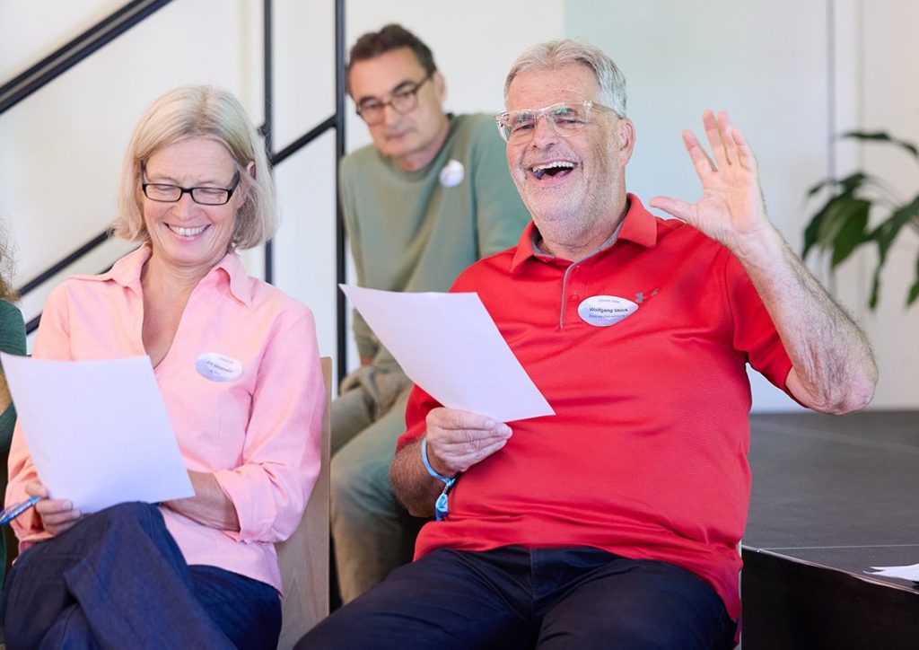 Eine Frau mit Brille und blonder Haarfarbe lächelt, während sie ein Blatt Papier hält. Neben ihr sitzt ein älterer Mann in einem roten Shirt, der fröhlich ist und seine Hand hebt. Im Hintergrund ist ein weiterer Mann mit Brille zu sehen, der ebenfalls ein Blatt Papier hat.