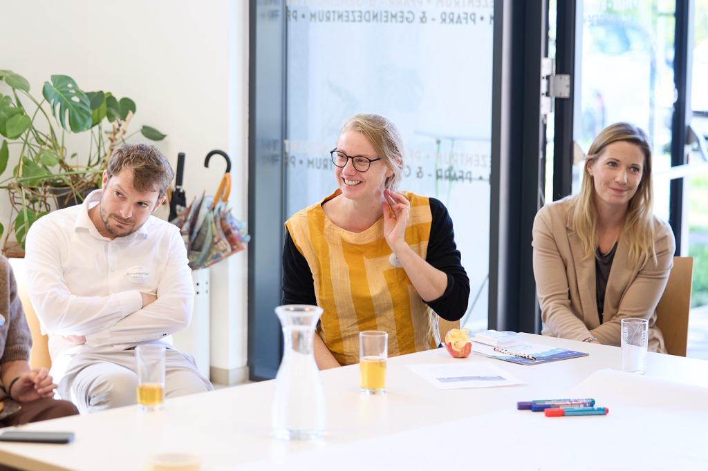 Eine Gruppe von Menschen sitzt an einem Tisch in einem hellen Raum. Eine Frau mit Brille und einem gestreiften Oberteil lächelt und spricht, während ein Mann in einem weißen Hemd aufmerksam zuhört. Im Hintergrund sind Pflanzen und verschiedene Gegenstände sichtbar.