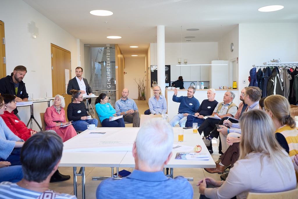 Eine Gruppe von Menschen in einem Raum, die an einer Diskussion teilnehmen. Sie sitzen um einen Tisch, während einige Personen sprechen und Notizen machen. Im Hintergrund ist ein Küchenbereich sichtbar.