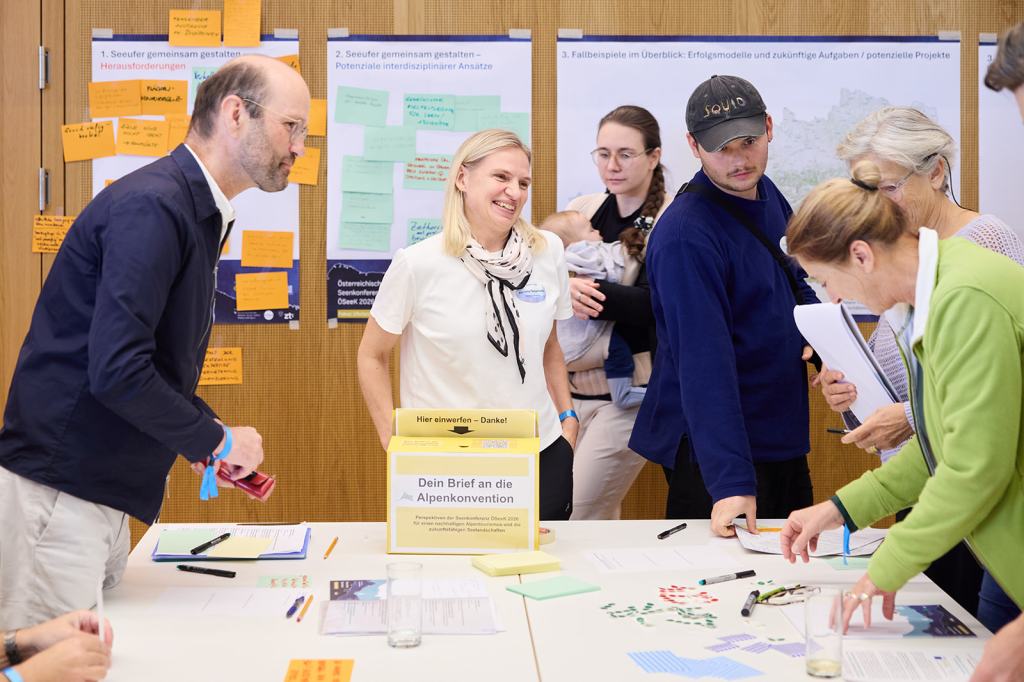 Eine Gruppe von Menschen diskutiert an einem Tisch, während sie an einem Workshop teilnehmen. Im Vordergrund steht eine Box mit der Aufschrift 'Dein Brief an die Alpenkonvention'.