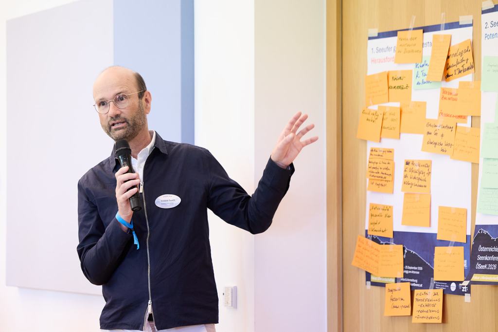 Ein Mann spricht in ein Mikrofon und präsentiert vor einer Pinnwand mit bunten Post-its während einer Konferenz.