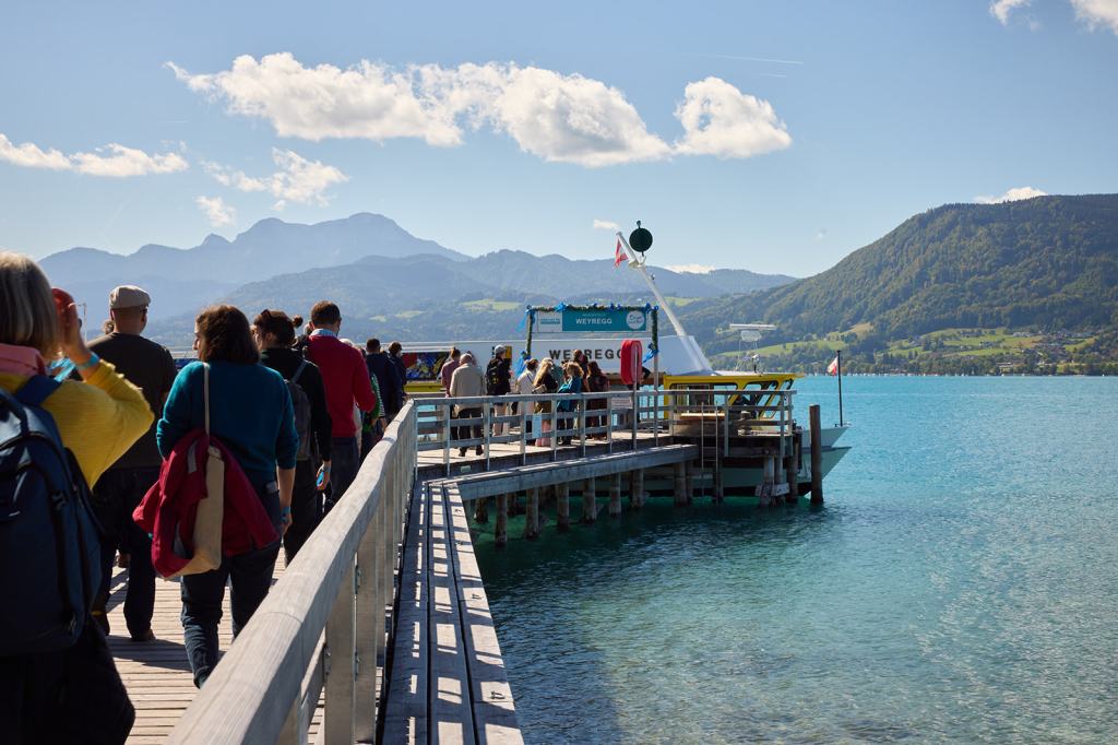 Menschen stehen auf einem Holzsteg am Wasser, mit Bergen im Hintergrund und einem Bootssteg, an dem ein Boot anlegt.