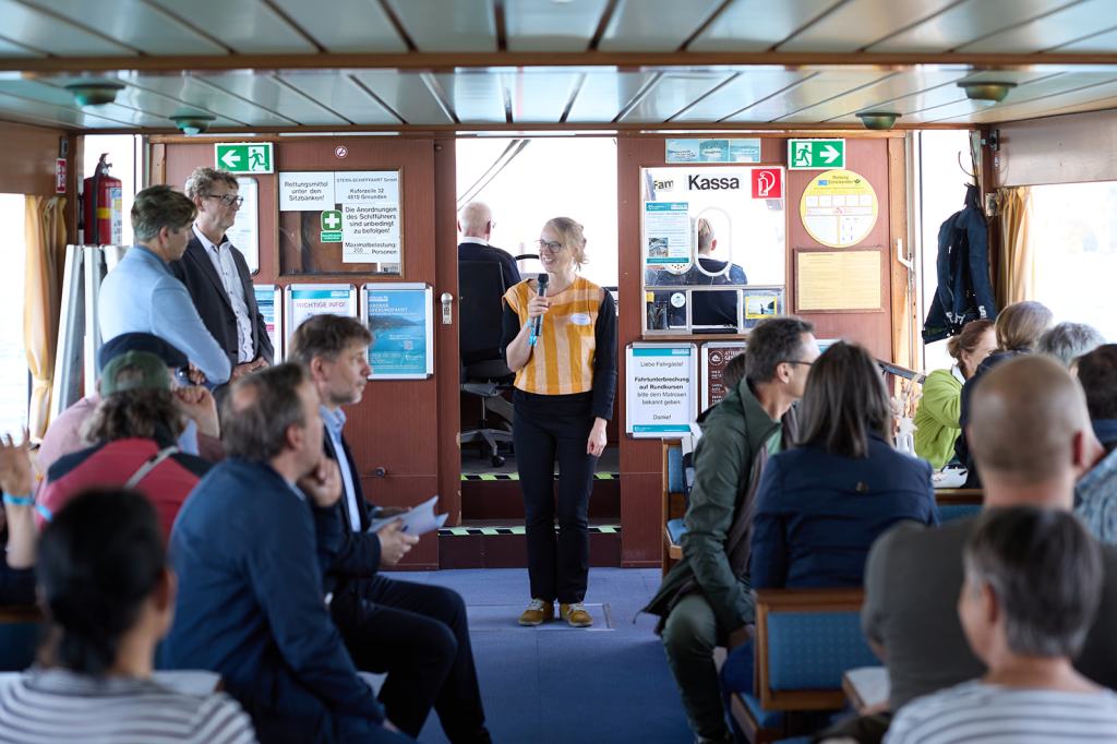 Eine Frau steht im Inneren eines Schiffes und spricht zu einer Gruppe von Menschen. Im Hintergrund sind weitere Personen und verschiedene Informationsschilder sichtbar.