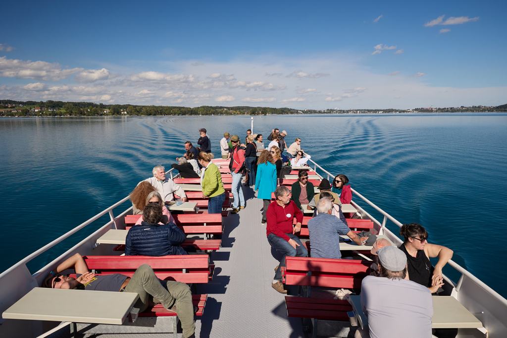 Eine Gruppe von Menschen sitzt und steht auf dem Deck eines Bootes, das auf ruhigem Wasser fährt. Im Hintergrund sind der Himmel, Wolken und eine Landschaft sichtbar.