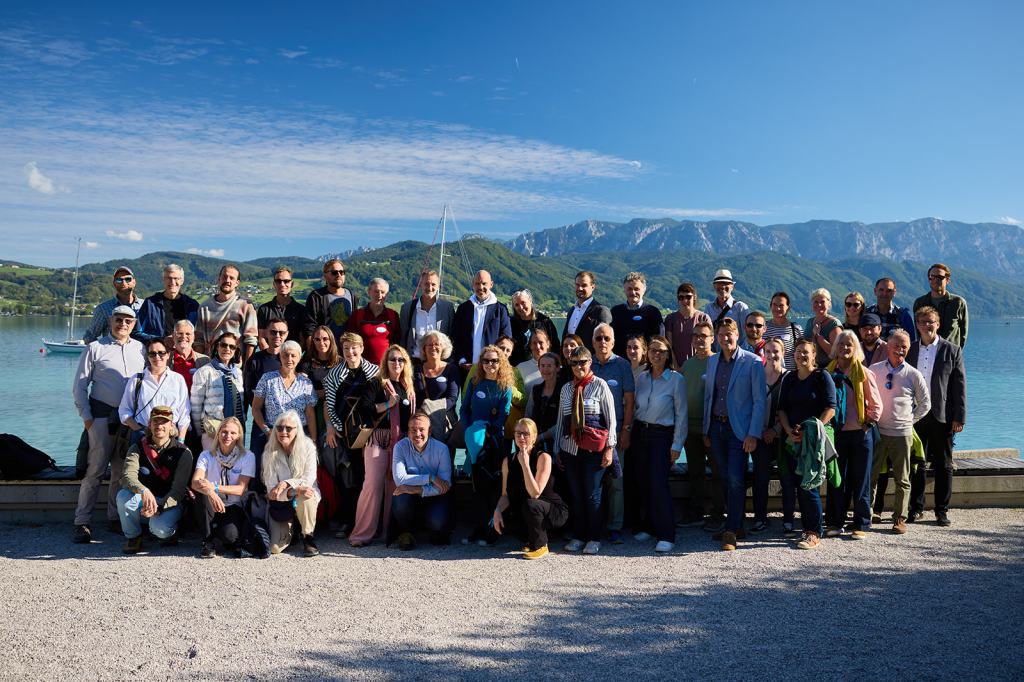 Eine große Gruppe von Menschen posiert am Ufer eines Sees mit Bergen im Hintergrund unter blauem Himmel.