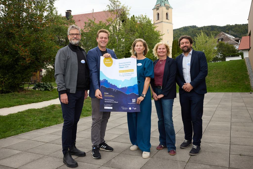 Fünf Personen stehen zusammen im Freien und halten ein Plakat für eine Veranstaltung. Im Hintergrund sind Bäume und eine Kirche sichtbar.