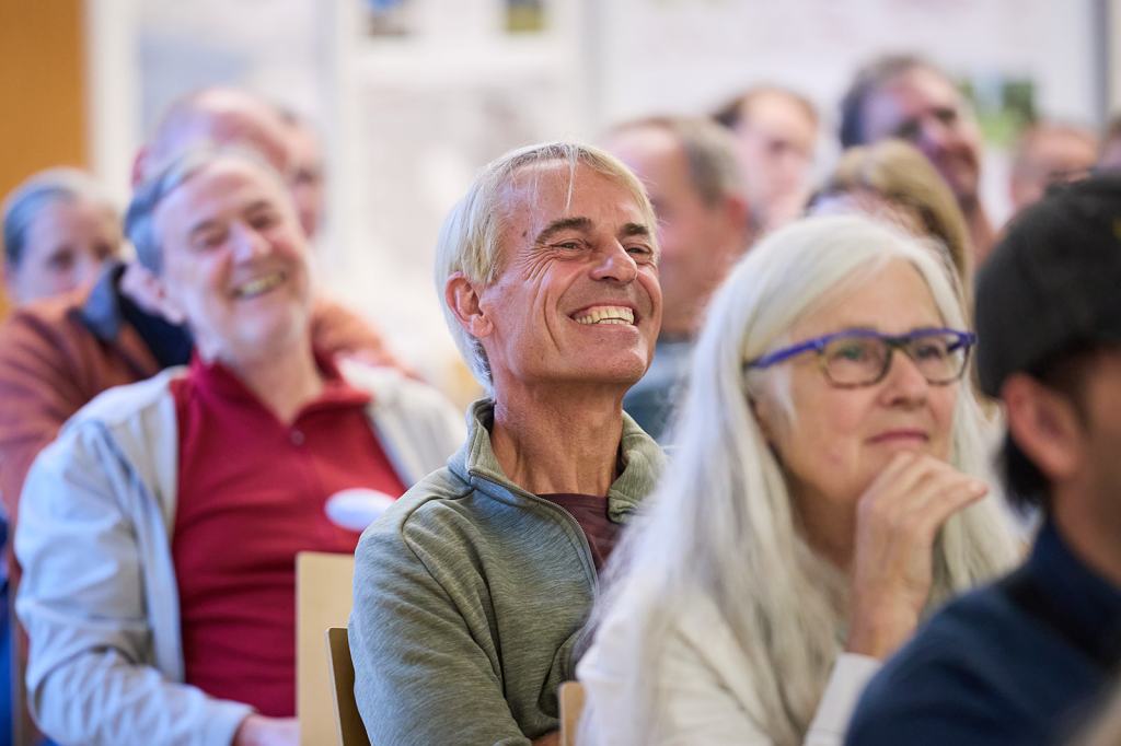 Eine Gruppe von Menschen, die in einem Vortrag sitzen und lächeln, während sie zuhören. Im Vordergrund sieht man einen Mann mit blondem Haar und einem breiten Lächeln sowie eine Frau mit grauen Haaren und einer Brille.