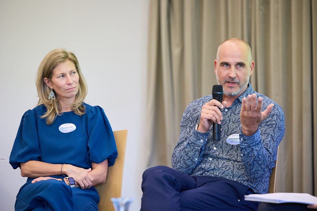 Ein Mann mit Bart spricht mit einem Mikrofon, während eine Frau mit lässiger Kleidung aufmerksam zuhört. Beide nehmen an einer Podiumsdiskussion teil.