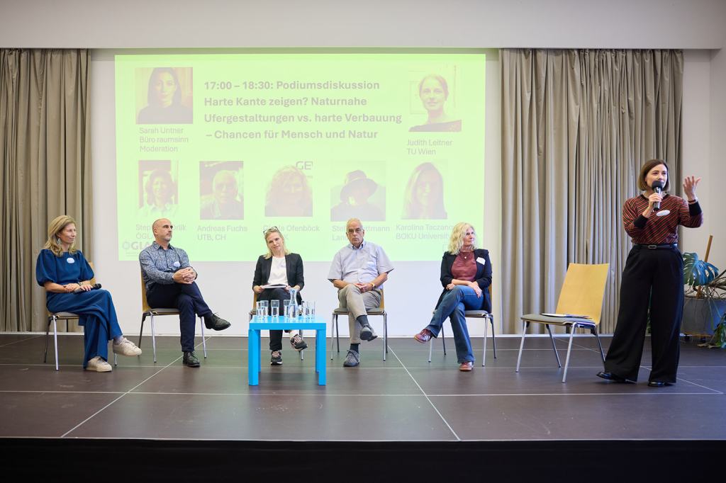 Podiumsdiskussion mit mehreren Personen, die über natürliche Ufergestaltungen und harte Verbauungen sprechen. Eine Moderatorin steht und spricht ins Mikrofon, während die anderen Teilnehmer auf Stühlen sitzen. Im Hintergrund sind Informationen zur Veranstaltung sichtbar.